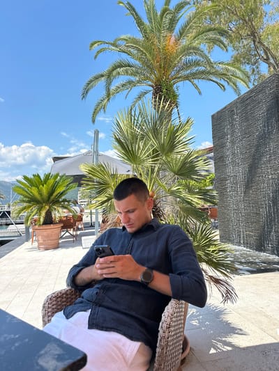 A young man sits in a woven chair outdoors, looking down at his smartphone. He is wearing a dark blue linen shirt and white pants. In the background, palm trees and a waterfall feature are visible, suggesting a tropical or Mediterranean setting. The man appears to be in a cafe or restaurant area, with tables and umbrellas also present. The sky is clear blue with a few scattered clouds. The location appears to be a waterfront area, possibly a marina, given the structures visible on the left side of the image. The overall impression is one of a relaxed, sunny day in a resort-like environment.