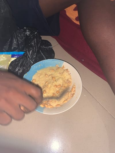 The scene, located indoors on a light grey tiled floor in Eziobodo, Nigeria, depicts a partial view of an individual. A light blue and white ceramic bowl containing a light-colored, viscous food substance, possibly a porridge, mixed with whole peanuts is centrally positioned. A portion of the food has been consumed. A dark-skinned hand is positioned over the bowl, appearing to scoop the contents. To the upper left of the bowl, several crumpled black plastic bags are present, partially obscuring a blue and white wrapper and clear plastic packaging. In the upper right, a dark-skinned bare leg rests on a red and orange patterned rug.