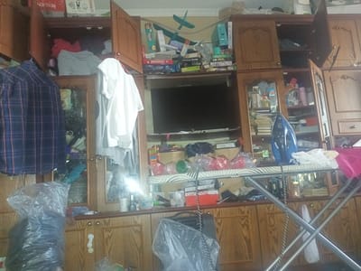 This is a medium shot, interior photograph of a wooden cabinet filled with various items, likely in a residential setting in Kamianske, Ukraine. The cabinet has multiple shelves and drawers, with some doors open and others closed.

On the upper shelves, there are board games such as Monopoly, as well as toys and other miscellaneous items. There is also a television set mounted in the center of the cabinet. Below the television, an ironing board is set up with an iron on it. There are also several plastic-wrapped items and bags, some containing what appear to be household goods.

The cabinet is well-stocked and somewhat cluttered, suggesting it's a functional storage unit. The lighting appears to be artificial, typical of an indoor environment. There is no indication of the time of day or weather outside. The overall impression is one of everyday domestic life.

Visible text includes "MONOPOLY" on a game box and "BOSCH" on a box of appliances. Other text is present on product packaging but is largely illegible.