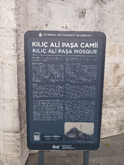 The image shows a black sign with white text attached to a white stone wall. The sign is located in Istanbul, Türkiye (latitude: 41.027, longitude: 28.98). The sign's text reads: "ISTANBUL BUYUKSEHIR BELEDIYESI KILIC ALI PASA CAMII KILIC ALI PASA MOSQUE." The sign features a QR code, two paragraphs of text, and a small black and white photograph at the bottom. The photograph depicts the mosque with a minaret, with people walking in front of it. There is a green bush in the bottom right corner. The sign is standing upright on a metal pole on a paved sidewalk.
