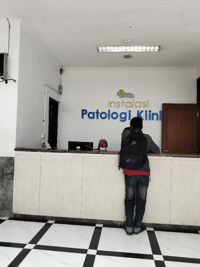 The image shows a man standing in front of a counter in a building located in Telukjambe, Indonesia. The man is wearing a black backpack and blue jeans, with a red shirt partially visible underneath. He is looking towards the right. The counter is white and has a sign that says "Instalasi Patologi Klin" above it, with a green logo of a cross with the word "Karawang" written in small letters underneath. Another person is partially visible at the counter behind the man, with a red mask on. The floor is black and white checkered tile. The room is lit by an overhead fluorescent light. There is a brown door in the background on the right side of the frame.