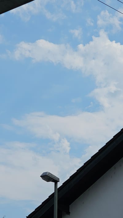 This is a low-angle outdoor photograph of a bright blue sky with scattered white clouds. A portion of a dark-colored roofline with an eave is visible in the upper right corner. Extending from under the eave is a metallic bracket holding a grey outdoor light fixture, which appears to be a floodlight. The fixture is angled downwards. In the upper left corner, a thin black wire or cable is visible against the sky. The scene suggests it is daytime, likely with fair weather given the clear sky and bright clouds. There is no text visible in the image. The perspective is from below, looking upwards towards the sky and the building's overhang.