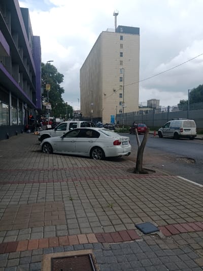 The image depicts an urban street scene in Newtown, Johannesburg, South Africa, specifically featuring the Premier Milling Silo (also known as Fatti's and Moni's building) in the background. The scene is captured during daylight under an overcast sky, viewed from a paved sidewalk.
**Foreground:** The sidewalk is constructed from grey and reddish-brown interlocking pavers. A metal manhole cover is visible in the bottom-left corner. Small patches of weeds emerge from cracks in the paving. A dead, Y-shaped tree trunk stands in a circular cutout in the pavement near the curb. An overturned black, rectangular electronic device (possibly a tablet) rests on the pavers to the right of the tree.
**Midground (Street and Vehicles):** A white BMW 3 Series sedan (E90 generation) is parked, with its right wheels on the street and left wheels on the sidewalk, facing away from the camera. Its rear license plate, partially visible, begins with "DZX". Behind it, a white Ford Ranger pickup truck is similarly parked. The asphalt road surface exhibits multiple large potholes and cracks. Further down the street, a red car is visible in motion, and a white delivery van with an orange traffic cone positioned behind it on the road is parked.
**Left Side:** A multi-story building with a purple-colored facade lines the left side of the street. Its ground floor features dark-framed shopfront windows. Several individuals are visible on the sidewalk in front of these shops; at least two are seated on the ground near bags of what appear to be goods or produce, suggesting informal vending. A black street light pole with multiple street signs and a "DHL" banner is present. Mature green trees are visible further down the street on this side.
**Right Side and Background:** The dominant feature on the right is the tall, rectangular, light-beige Premier Milling Silo, exhibiting a vertically ribbed texture and irregularly spaced small windows. A dark structure with an antenna-like mast is present on its roof. In front of the silo, a long, corrugated grey metal fence runs parallel to the street. Another multi-story building is partially visible in the far background beyond the fence.