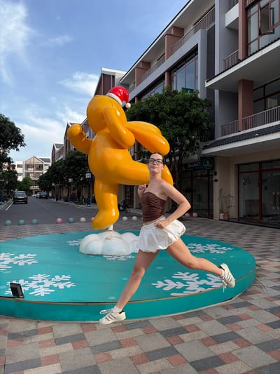 An eye-level medium shot captures a young woman with glasses mid-jump in front of a large, bright orange, inflatable sculpture of a cartoon character wearing a Santa hat. The sculpture is positioned on a teal-colored circular platform decorated with white snowflakes. The woman is wearing a brown strapless top and a white ruffled skirt, with white sneakers. Her body is angled towards the right, with her left leg extended forward and her right leg bent. The background reveals a street scene in Nghĩa Trụ Commune, Vietnam, with modern buildings lining the road. The buildings are multi-story with a mix of beige, grey, and brown facades, featuring balconies and large windows. Trees are planted along the sidewalk. Cars, motorcycles, and decorative spherical bollards are visible on the street. The sky is partly cloudy with a bright blue hue. The ground is paved with a checkered pattern of grey, pink, and beige tiles. The overall scene appears to be an outdoor public space, possibly a plaza or a pedestrian area.