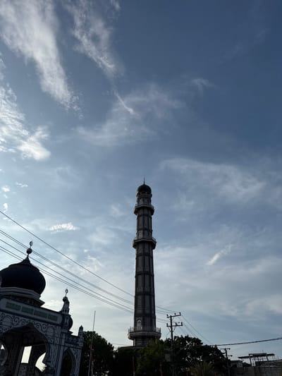 In Keudekruenggeukueh, Indonesia, the image captures the imposing minaret of Masjid Besar Bujang Salih against a vast, partly cloudy sky. The minaret, a tall, octagonal structure with multiple levels and decorative elements, dominates the right side of the frame. To the left, a portion of the mosque's entrance is visible, featuring a domed roof and an arched gateway with intricate patterns. Several power lines traverse the foreground, running horizontally across the scene. Lush green trees are visible behind the mosque and minaret. The overall atmosphere is calm and serene, with the architecture of the mosque standing as a prominent landmark within the urban landscape.