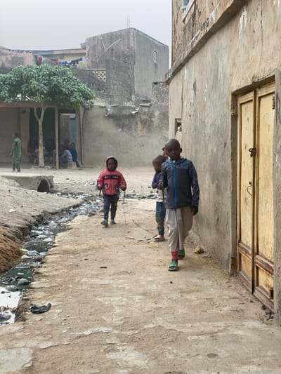 Three children stand near a trench filled with refuse. The boy in front wears a red jacket and holds a string. Two other boys are behind him; one has a blue jacket, the other wears purple. Nearby, a group of people sits under a leafy tree. A worn building looms in the background, its walls aged and weathered, and a small door is visible above. The ground appears cracked and uneven.