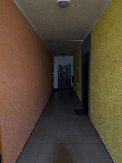 The image shows a long, narrow hallway with contrasting walls of orange on the left and yellow on the right, separated by a dark gray section. The ceiling is a light gray with a circular light fixture. The floor is tiled with dark gray square tiles.
At the far end of the hallway, there are two doors. The door on the left is dark gray with a numbered plate that reads "78". To the right of this door, a dark-colored electric scooter is parked. The door on the right is black with a silver handle. A small dark doormat is placed in front of this door.
The hallway appears to be located in a residential building, likely an apartment building entryway or corridor. The architecture and color choices are typical of many buildings in Irpin, Ukraine. The overall scene is clean and appears well-maintained, with no signs of damage or conflict. The lighting is dim, suggesting it may be evening or the lights are not fully illuminated. There are no people visible in the image, and no actions or interactions are taking place.
