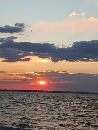 The image depicts a sunset over a body of water, likely the Black Sea, near Zatoka, Ukraine. The sun, a bright orb partially obscured by clouds, is low on the horizon, casting a warm glow of orange and red across the sky and water. Dark, dramatic clouds streak across the upper portion of the sky, contrasting with softer, pink-tinged clouds nearer the horizon. The water is choppy, with visible ripples and waves. A dark, silhouetted shoreline with trees is visible in the distance, separating the water from the sky. The overall atmosphere is serene and picturesque, characteristic of a coastal sunset.