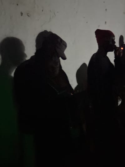 The image shows a dimly lit scene with several silhouetted figures against a textured, off-white wall. The lighting appears to be from the side, casting distinct shadows. In the foreground, on the right side of the image, a person wearing a red beanie is visible in silhouette, with their head tilted as if smoking a cigarette, indicated by a small, bright red light near their mouth. To the left of this figure, another person is also silhouetted, their head obscured by a cap or hood. Further to the left, a larger, more defined shadow of a person is visible, seemingly in profile. The environment appears to be indoors or a covered outdoor space with a rough-textured wall, possibly concrete or stucco, exhibiting some imperfections. The lower left portion of the image has a greenish cast, possibly from a light source. The overall impression is a candid, low-light moment captured in an urban setting within Wumat, Nigeria.