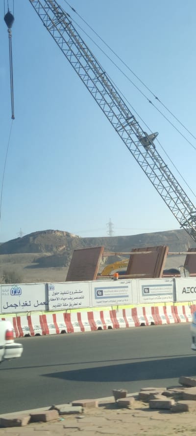 The image shows a construction site in Jeddah, Saudi Arabia. A large crane dominates the right side of the frame against a clear blue sky. In the background, there is a low mountain range with electricity pylons. A yellow excavator is visible behind a set of safety barriers, which have signs with Arabic text, the logos of "Amanat Jeddah," and the text "AECO." The signs also mention a project to improve rainwater drainage for a more beautiful Jeddah, referencing "the old Mecca road." Parts of two cars are visible on either side of the foreground. The ground on the roadside is scattered with bricks.