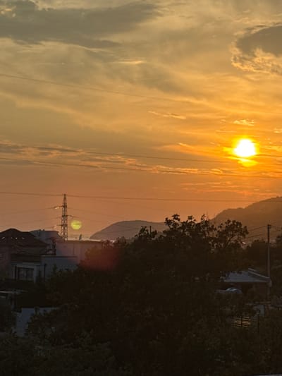 The image captures a sunset over a residential area with a view of hills and the sea in the distance. The sky is filled with vibrant orange and yellow hues, with scattered clouds. A bright sun is visible near the top right corner. In the foreground, silhouetted trees and the rooftops of houses are visible. A power line tower stands prominently in the mid-ground, with some electrical wires crisscrossing the scene. The overall impression is one of a serene evening in a coastal town. The image is an outdoor, daytime shot with no people visible. The location is Sustaš, Montenegro.