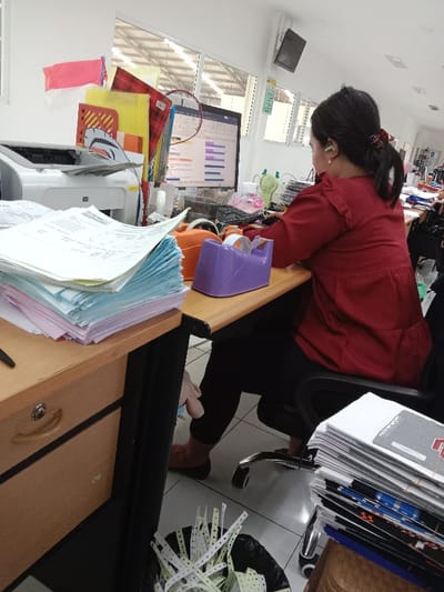 A woman in a red shirt is seated at a desk, using a computer in an office located in Kecamatan Ngaliyan, Indonesia. The woman's legs are crossed and she is using her right hand to manipulate the mouse on the desk. There are a large stack of papers on the desk in front of her. A printer is located on the desk to the left of the stacks of paper. A tape dispenser, a pair of scissors, and a pair of headphones are also present on the desk. The woman is seated on a black chair, with a black chair next to her, also occupied. This chair has a large stack of papers on its seat. On the floor, there is a trash can filled with discarded paper. The office appears to have a light-colored tiled floor, white walls, and windows overlooking an industrial area.