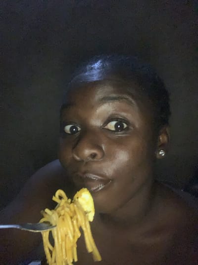 In the city of Agban, Nigeria, a young Black woman with dark skin and short hair is captured in a dimly lit, close-up selfie. She is looking directly at the camera with wide, curious eyes, her mouth slightly open as she brings a forkful of spaghetti to her lips. The spaghetti appears to be cooked and coated in a sauce, with a piece of what looks like scrambled egg on the fork as well. She is wearing a small, sparkling stud earring in her visible ear. The background is dark and indistinct, suggesting an indoor setting at night. The lighting is minimal, highlighting her face and the food.
