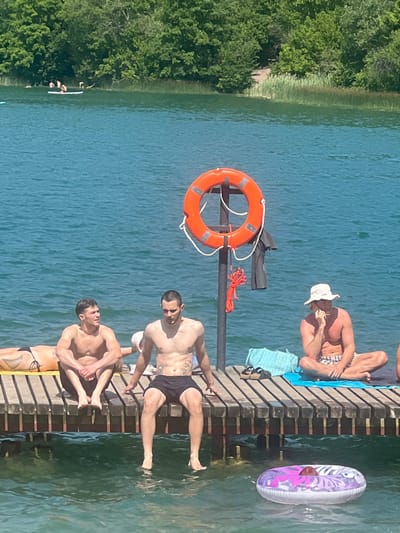 The image shows several people enjoying a sunny day at a lake or river in Vilnius, Lithuania. The scene is set on a wooden dock extending into the clear blue water. Lush green trees line the shore in the background.
On the dock, four men are visible. One man, wearing a white hat and checkered shorts, is sitting down. Another man, in black shorts, is standing and looking towards the camera. Two other men are sitting or lounging on the dock. One of them appears to be wearing black swim trunks and the other is wearing bikini-style bottoms and fishnet stockings. In the water next to the dock, a colorful inflatable ring floats. A life preserver ring is mounted on a pole on the dock.
The time of day appears to be midday, given the bright sunlight casting shadows. The weather seems to be warm and clear, ideal for swimming and relaxing by the water. The overall mood is one of leisure and enjoyment. There is no visible text in the image.