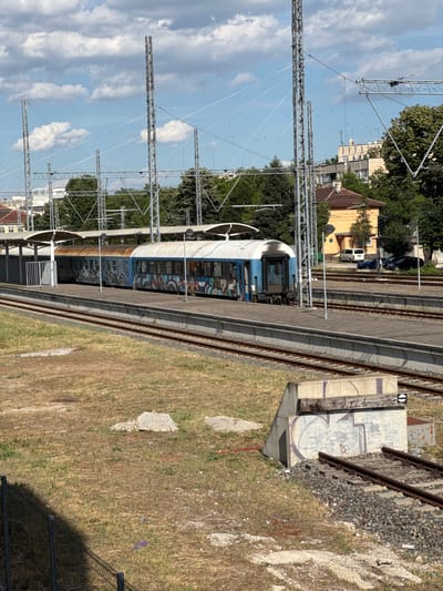 This vertically oriented image depicts a train station scene in Vidin, Bulgaria, under a bright, sunny sky with scattered clouds. The primary subjects are a blue and white passenger train covered in graffiti, situated on railway tracks at a station platform. The train's carriages show signs of graffiti art.
The scene includes the railway infrastructure, such as overhead power lines supported by metal pylons, and the tracks themselves, ballasted with gravel. A concrete structure with graffiti is visible next to the tracks, possibly part of the station's signaling system. Adjacent to the platform, dry, yellowish grass covers the ground, with some scattered concrete debris and a patch of red soil.
In the background, trees and buildings are visible. A yellow building with a reddish-brown roof and a larger, multi-story building with a light-colored facade suggest an urban or semi-urban environment. The time of day appears to be midday, given the strong sunlight and shadows. There are no visible people in the image. The overall impression is of a functional, somewhat weathered, European train station on a clear day.