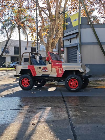 The image depicts a red and beige Jeep Wrangler, customized with "Jurassic Park" decals and the number 12, parked on a city street. The Jeep features red wheels with BFGoodrich Mud-Terrain T/A tires. The street shows signs of wetness and reflections. In the background, a building with "Western Union" and "Pago Facil" signage is visible. Trees with autumn foliage are also present. The location is likely La Plata, Argentina. There are faint silhouettes of individuals in the background of the image.