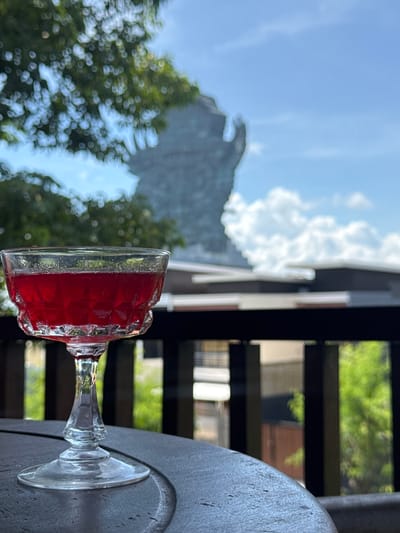 A tilted, eye-level shot features a glass of red liquid on a gray, round table. Outside the balcony, which has wooden railings, are lush, green trees, a building with gray siding, and a blue sky with white clouds. The scene is located in Ungasan, Indonesia.
