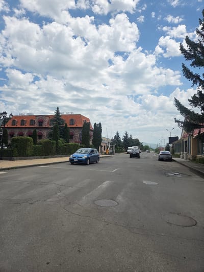 The image captures a street scene in Gyumri, Armenia. In the foreground, a blue hatchback car is driving on a paved road with a pedestrian crossing. The road is lined with sidewalks and some greenery. A multi-story building with a red tiled roof, identified as "GYANHOTEL" by signage, is visible on the left side of the street. The building features arched windows with red awnings. Several trees and manicured bushes are planted in front of the hotel. In the background, other cars and buildings can be seen, and mountains are visible in the distance under a sky with fluffy white clouds. The scene appears to be during the daytime.