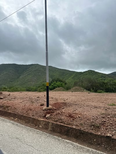 This image depicts a scenic view of a mountainside in La Asuncion, Venezuela. The scene is dominated by lush green, tree-covered hills that stretch across the frame under a cloudy, overcast sky. The terrain in the foreground consists of a rough, reddish-brown dirt path and a paved road, separated by a low, weathered curb. A vertical metal pole, partially painted with black and yellow bands, stands in the foreground, with a thin wire extending from its top. The overall atmosphere suggests a tranquil, possibly humid, day in a mountainous region. There is no visible text in the image.