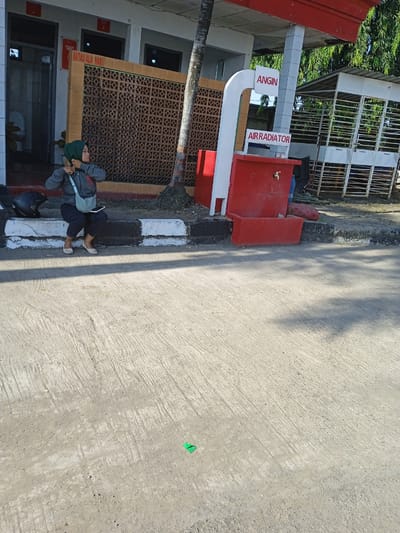 A woman wearing a hijab and casual clothing sits on a black and white curb adjacent to a building in Brangsong, Indonesia. She adjusts her jacket, with a bag slung over her shoulder and a helmet placed on the curb beside her. To the right of the woman is a sign reading "ANGIN AIR RADIATOR" atop a red structure with a faucet. The building behind her has a patterned brick wall and signage. A tree trunk is situated next to the sign, and a metal structure is visible in the background. The foreground is dominated by a concrete surface.