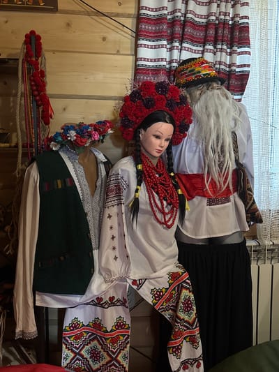 In Chorna Tysa, Ukraine, a display of traditional Ukrainian folk attire is presented. Two mannequins are featured: one female, adorned with a large, circular headdress of red flowers and intricate beadwork, a white embroidered blouse, and patterned trousers. The second, a male figure, wears a white embroidered shirt, a red sash, a richly decorated hat, and a long white beard. A third, partially visible mannequin on the left wears a green vest over a light-colored shirt and a colorful floral headdress. The background consists of wooden paneled walls, red and white patterned curtains, and a white lacy curtain. Decorative items, including ropes with red pompoms and strands of beads, hang on the left. The overall scene appears to be an indoor exhibition or a shop display showcasing cultural heritage.