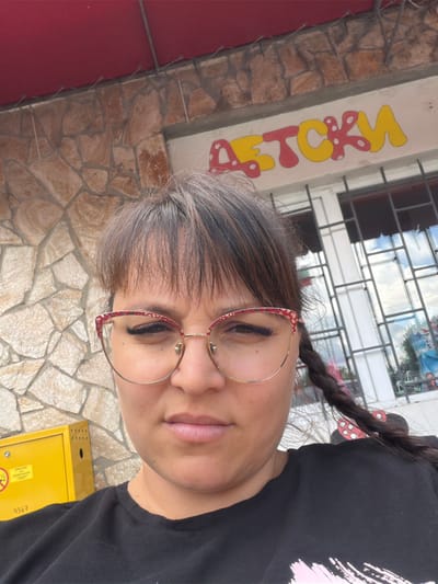 A woman with brown hair and glasses poses for a selfie outdoors. She is wearing a black t-shirt and is looking at the camera with a neutral expression. Behind her, a stone wall is visible, and above that, a red awning. To her right, a sign with yellow and red lettering that appears to read "ДЕТСКИ" (DETSKI, meaning children's in Russian) is mounted on a building. A yellow metal box with some markings and the number "4367" is to her left. The location is identified as Ravno pole, Bulgaria.
