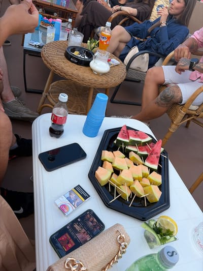 In a relaxed outdoor setting, likely on a balcony or patio in Troyanovo, Bulgaria, a group of people are gathered around a table laden with refreshments. The focus is on a white table with various items arranged on it. In the foreground, a black phone rests on the table, accompanied by a pack of "Ome" cigarettes. Further into the scene, a black plastic tray holds an assortment of fruit skewers, featuring watermelon and cantaloupe. Nearby, a small glass container holds sliced cucumber, lemon, and parsley. A bottle of Coca-Cola and a blue plastic cup are also present. In the background, a woman in a denim dress is seated, smoking, while other individuals are partially visible, suggesting a social gathering. The atmosphere appears casual and leisurely.