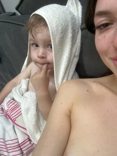 A close-up, shoulder-level selfie captures a woman on the right and a toddler on the left, both seemingly at home in Эстачи, Russia. The woman, with her face partially visible and her shoulder exposed, is looking into the camera with a slight smile. The toddler, wrapped in a white hooded towel with pink stripes underneath, is positioned to the left of the woman. The toddler's eyes are open, and they are looking to the side with one finger in their mouth. The background consists of a dark, plush surface, possibly a sofa. The lighting appears to be natural, creating a soft ambiance.