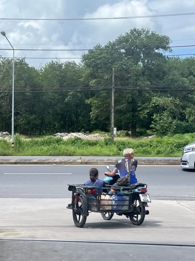 Two people are on a three-wheeled motorcycle with a sidecar. The driver, a person with white hair and sunglasses, wears a blue and black cycling jersey. A passenger with dark hair pulled into a ponytail, wearing a blue shirt, sits in the sidecar. The motorcycle has a license plate with Thai characters and the number "555". They are on a paved road next to a grassy median with trees and a pile of debris in the background. A white car is partially visible on the right. The scene is in Pa Khlok, Thailand, with a lamppost and power lines overhead.