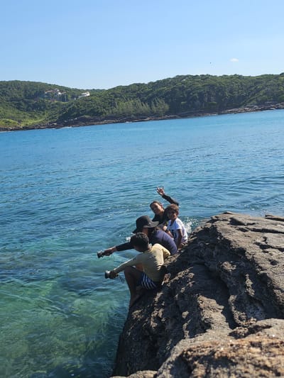Four individuals are positioned on a rocky outcrop adjacent to the ocean. The rocky surface is dark in color. The ocean exhibits a turquoise tint near the rocks, transitioning to a deeper blue further out. One person is pointing to an object beyond the frame. A coastline covered in lush greenery stretches into the distance. Two people are holding cylinder objects. The location is Armação dos Búzios, Brazil.