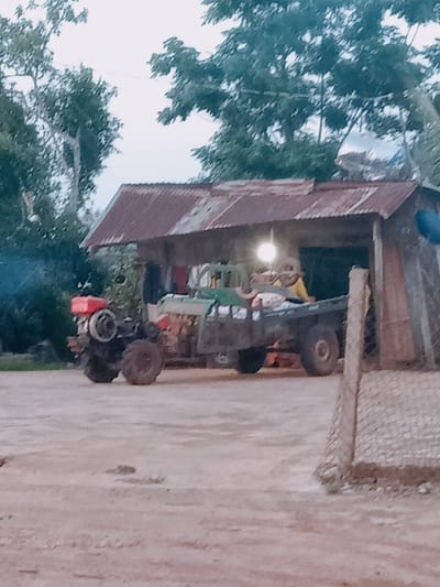 The scene appears to be a rural or semi-rural location in Xuan Long, Vietnam. There is a makeshift structure with a corrugated metal roof, likely serving as a shelter or open-air garage. Inside the structure is a small tractor with a trailer. The trailer contains people. The ground in front of the structure is dirt. Trees are visible in the background. A section of what appears to be a chain-link fence is visible in the lower right corner.