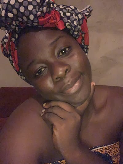 The image shows a close-up, slightly grainy, self-portrait of a Black woman wearing a headscarf. She is looking directly at the camera with her chin resting on her hand. Her headscarf has a dark blue, white, and red polka dot and floral pattern. She is wearing a patterned garment around her shoulders and upper chest, with a colorful band visible at the bottom of the frame. The background appears to be a plain, textured wall. The lighting is dim, creating a warm glow on her skin. The woman's features are prominent, with her eyes open and a slight smile on her lips. Her skin appears smooth and dewy. The overall impression is intimate and relaxed. The image was taken in Agban, Nigeria.