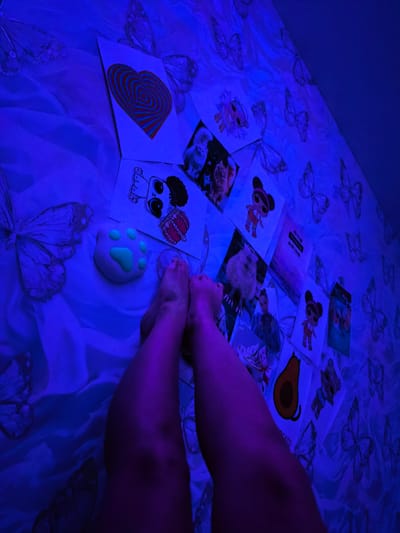 A person's legs are visible in the foreground of the image, bent and resting on a bed with a butterfly-patterned sheet. The sheet is illuminated by blue light. Scattered on the bed are several printed images, including a heart-shaped drawing, cartoon characters, an avocado, and images of animals. A light-colored paw-shaped object with green pads is also present on the bed. The scene appears to be an indoor setting, possibly a bedroom. There is no specific indication of the city of Tver, Russia.
