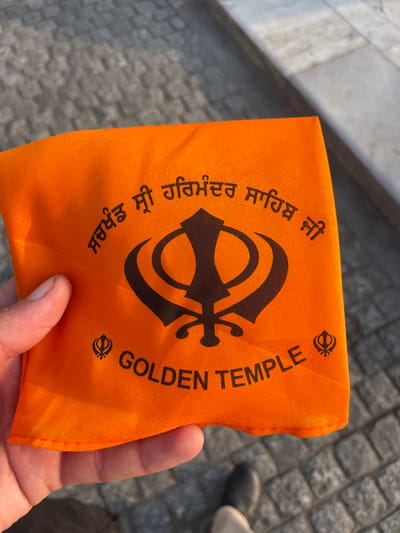 An orange cloth with black lettering and a symbol is being held by a person's hand. The cloth displays the Sikh Khanda symbol and the words "GOLDEN TEMPLE" in English. Above the symbol, in Punjabi script, are the words "ਸਰਬੱਤ ਦਾ ਭਲਾ" (Sarbat da Bhala), which translates to "Good for all". This cloth is likely a souvenir or religious item from the Golden Temple (Harmandir Sahib) in Amritsar, India. The background shows a stone pathway and what appears to be part of a large marble structure, possibly a wall or staircase, also consistent with the architecture found at the Golden Temple complex.
