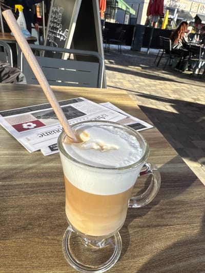 The image presents an outdoor cafe scene in Reichstett, France, viewed from a low perspective. In the immediate foreground, a clear glass mug, featuring a handle and a footed base, contains a three-layered beverage. The liquid layers are dark brown at the bottom, transitioning to a lighter brown in the middle, topped by a thick stratum of white foam. A light-colored, possibly paper, straw is inserted into the foam at an acute angle. The mug rests on a light-brown wooden table exhibiting distinct grain patterns.

Behind the beverage, a folded newspaper or magazine, partially readable as "L'Ancienne," lies flat on the table. The mid-ground reveals an outdoor patio paved with patterned stones, illuminated by direct sunlight, casting sharp shadows. Several metallic grey outdoor tables and chairs are visible. To the left, a black A-frame chalkboard sign displays white text. In the background, two individuals are seated at a table; one person with long, reddish-brown hair is discernible. Another person's back is partially visible to the far left. Distant elements include parts of buildings and red and green awnings or umbrellas.
