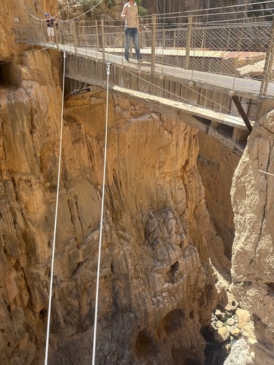 The image depicts the Caminito del Rey, a pathway built along the steep walls of the Gaitanes Gorge in El Chorro, near Álora, in the province of Málaga, Spain, which is part of the larger municipality of Antequera. In the foreground, a suspension bridge made of metal and wood spans a deep gorge. Two individuals are visible on the bridge. One, wearing a blue helmet, shorts, and a checkered shirt, is standing in the middle of the bridge, holding a camera. Another person, wearing a light-colored t-shirt and jeans, is further along the bridge, walking away from the camera. The bridge is suspended by thick white ropes anchored to the sheer, rocky cliff faces that rise on either side of the gorge. The rock formations are rugged and appear to be composed of layered sandstone, with deep crevices and weathered surfaces. The gorge itself is deep and narrow, with a visible rocky bottom and some water at the very bottom of the frame. The sunlight casts strong shadows, highlighting the dramatic topography of the gorge and the structural elements of the bridge. The environment is natural and rugged, characterized by the imposing scale of the cliffs and the precarious nature of the walkway.
