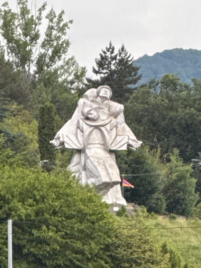 This is a landscape image featuring a large, light-colored stone statue of a person, likely religious in nature, positioned outdoors. The statue is oriented diagonally in the frame, with its head towards the bottom left and feet towards the top right, creating a dynamic perspective. The figure appears to be draped in flowing robes.

The statue is situated amidst lush green foliage, suggesting a natural setting with trees and hills. In the background, a misty or cloudy sky is visible, with hints of blue suggesting daylight, possibly morning or afternoon. The overall atmosphere seems serene and contemplative, likely a tourist or spiritual site. A small, colorful flag or banner, featuring orange and blue sections, is visible on the right side of the statue, near its feet. A thin wire or cable stretches across the frame, passing behind the statue. The image has a somewhat grainy texture, typical of older or lower-resolution digital photography. The location is identified as Dilijan, Armenia, which is known for its natural beauty and historical sites.