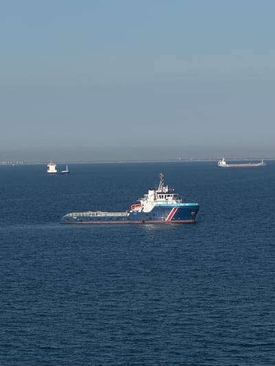 The image depicts a wide, calm expanse of deep blue ocean under a hazy, light blue sky. In the foreground, a modern blue and white vessel with red accents is moving across the water. The vessel has a multi-level superstructure with windows, a radar, and antennae. The hull is detailed with a blue stripe and a red waterline. In the middle ground, a smaller white and blue vessel is also visible. Further in the distance, a long, light-colored cargo ship with a pink stripe along its deck is on the horizon, as is a faint landmass with a cluster of structures. The overall scene is serene and industrial, suggesting maritime activity. There is no indication of the city of Unknown, Italy, in the image.