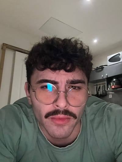 A person with curly dark hair and glasses poses for a selfie. The image is taken from a low angle, showing the person's face and upper torso. They wear a sage green shirt and sport a fake mustache. In the background, there is a countertop with kitchen utensils, some bottles, and a white sound system. The room is well lit.