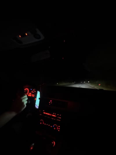 The image depicts a nighttime scene from the interior of a vehicle being driven on a road in Goritsa, Bulgaria. The perspective is from the driver's or front passenger's seat, looking forward through the windshield.

**Car Interior:**
The car's interior is largely dark, illuminated by various red and some white/blue dashboard lights.
- A person's left hand and forearm are visible on the left side of the frame, gripping a dark steering wheel at approximately the 9 o'clock position, indicating the driver's presence.
- The dashboard directly in front of the driver displays red-illuminated analog gauges, including a speedometer showing a speed between 100 and 120 units (likely km/h). Other indicator lights are present, some appearing yellow or orange.
- To the right of the steering wheel, a mobile phone is mounted, its screen brightly illuminated with a blue-green interface, displaying what appears to be a navigation or map application.
- The central console features red-illuminated controls for the audio system and climate control. The radio display shows "FRH 02:27" and "21:50," possibly indicating a radio frequency and time.
- An overhead console, visible in the upper portion of the frame, has two small red lights illuminated.
- The overall aesthetic of the interior lighting and dashboard design suggests a BMW vehicle of a certain generation (e.g., E8x/E9x series).

**Exterior/Environment:**
- The vehicle is traveling on a dark, paved road, illuminated by its headlights. White lane markings are visible on the asphalt.
- In the distance, multiple sets of white headlights are visible, indicating oncoming traffic. Further ahead on the right side of the road, red taillights or reflective markers are discernible.
- The roadside on both sides appears dark, likely consisting of vegetation. A distinct red reflective marker is visible on the right shoulder of the road.
- The sky overhead is completely dark, confirming nighttime conditions.