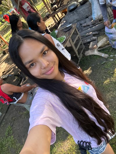 The image depicts an outdoor scene in what appears to be an informal gathering or communal cooking area, specified to be in Carmen, Philippines. The ground is a mix of dirt and sparse green grass, illuminated by natural daylight.

In the immediate foreground, a young individual, presumably female, with long, dark, straight hair, smiles directly into the camera, taking a selfie. She wears a light purple short-sleeved t-shirt with a multi-colored emblem visible on the left chest area, and light-wash denim shorts. A black claw hair clip is attached to her shorts on the lower right side.

In the midground, several individuals are present. To the left, a person with dark hair and a green top looks towards the right. Behind them, another individual with dark hair wears a white sleeveless top and a hoop earring, also looking right. Further left and towards the bottom, a person wearing a red and black tank top is seated, facing away from the camera.

In the background, on the right side of the frame, a person with a striped long-sleeved shirt is partially visible, holding a patterned bag containing large orange produce, possibly papayas.

The central background features a makeshift cooking and preparation area. A rustic wooden table or bench holds a white plate containing chopped green vegetables, possibly a salad, and a knife. Behind this table, an outdoor cooking setup is visible, featuring various dark metal pots and pans, suggesting use over an open fire or simple stove. Logs, wooden planks, and other debris are scattered around the area, contributing to the informal nature of the setting.