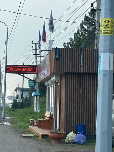 The image shows a street scene in Listvyanka, Russia. A red LED sign displaying "CIRCUM-BAIKAL" is prominently displayed on a metal pole. To the right of the sign is a wooden-paneled building with large windows. Several flags are visible on poles in front of the building, including the Russian flag and other national or organizational flags. A utility pole stands in the foreground on the right, with yellow flyers attached, one of which displays phone numbers for taxi services. The ground is paved on the left, with a wet patch and some debris, and a grassy area with potted plants and a wooden bench on the right. In the background, trees and buildings can be seen under an overcast sky. Trash bags and a blue trash bin are located near the building.
