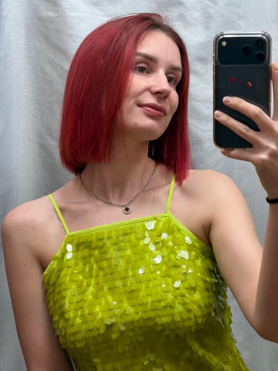 A woman with auburn hair is holding a smartphone, presumably taking a selfie. She is wearing a lime green, sequined top and a silver necklace. The background appears to be a white sheet. The location is not discernible from the image.