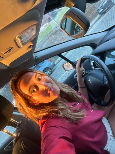 The image captures a young woman seated in the driver's seat of a car, illuminated by warm, golden sunlight characteristic of late afternoon or early evening. She has long, light brown hair that catches the light and is wearing a maroon or dark pink collared shirt. Her face is turned towards the camera, displaying a calm expression and a slight smile, with her eyes looking forward.
Her right hand rests on the steering wheel, which prominently features a Peugeot lion emblem. The car's interior is visible, including parts of the dashboard, a rearview mirror, and an overhead interior light fixture. Through the front windshield and reflected in the side mirror, other cars are visible on a paved surface, suggesting the car is parked on a street or in a parking lot. The rearview mirror also shows reflections of another vehicle and a building.
The scene conveys a sense of peacefulness, highlighted by the soft, directional sunlight casting warm tones on the woman's face and hair. The setting is identified as being within Aldoar, Foz do Douro e Nevogilde, Portugal.