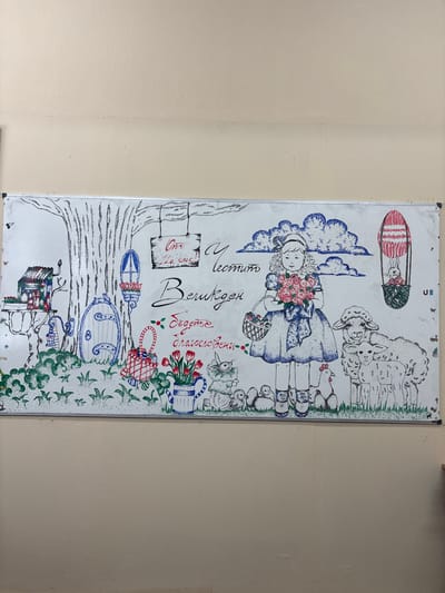 A rectangular whiteboard, measuring approximately 1.5 meters by 0.75 meters, is mounted on a light beige, textured wall in a room in Cherven Bryag, Bulgaria. The whiteboard features a detailed, multi-colored drawing executed primarily with marker pens and pointillism techniques, depicting an Easter or spring-themed scene.

The drawing portrays a landscape with a large, bare tree on the left, from which a small, red-roofed house is suspended. A round blue door with white detailing is situated at the tree's base, and a small, plant-filled lantern hangs nearby. A sign reading "От 1a клас!" (From 1a class!) in red and black text hangs from a tree branch. In the foreground, green dotted foliage covers the ground. A striped red and white basket sits on the ground, alongside a blue and white striped pitcher containing red tulips.

To the right, a young girl with blonde hair, dressed in a blue, dotted dress and a bow, holds a bouquet of pink roses and a small basket. Adjacent to her, a mother sheep and a lamb are depicted. Near the girl's feet are several small chicks, one chicken, and a rabbit statue. Above, blue dotted clouds float in the sky. To the far right, a red and white hot air balloon, styled as an Easter egg, carries a small rabbit in its basket. Large script in black reads "Честит Великден" (Happy Easter), with "Бъдете благословени" (Be blessed) written below in red script. The whiteboard itself has a silver metallic frame and shows minor scuff marks.