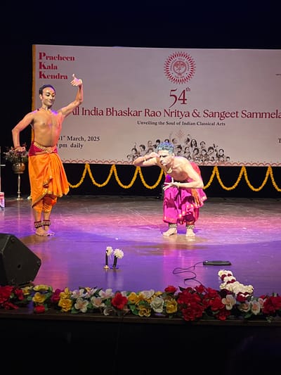 The image depicts a stage performance of Indian classical dance. Two dancers, one male and one female, are performing on a stage. The stage is decorated with garlands of colorful flowers. The male dancer is wearing a bright pink and yellow costume, while the female dancer is wearing a bright orange and red costume. Both dancers are barefoot. The male dancer is kneeling and extending his left hand towards the audience.  A banner behind the dancers reads "Pracheen Kala Kendra" and "54th All India Bhaskar Rao Nritya & Sangeet Sammela Unveiling the Soul of Indian Classical Arts". The performance appears to be taking place at Pracheen Kala Kendra, a cultural center located at the given coordinates (latitude: 30.733, longitude: 76.789), in Sector 35, Chandigarh, India. The floor of the stage is polished wood. A microphone stand and a few flowers can be seen at the front of the stage.  The stage is illuminated by spotlights.  The background is dark.  The image appears to be a still from a video recording of the performance.