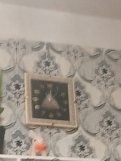 The media file shows a framed square clock on a wall with a repeating damask-style wallpaper pattern in shades of grey and white. The clock face is dark with Roman numerals and visible hands, suggesting it is a functional timepiece. A small, light-colored figurine with an orange nose is partially visible in the lower portion of the image, peeking out from behind the clock. The overall impression is of a domestic interior setting, possibly a living room or hallway, given the clock on the wall and the decorative wallpaper. There is no information about the time of day, weather, or specific emotions conveyed. No visible text is present in the image. The context provided suggests this scene is located in Termez, Uzbekistan.