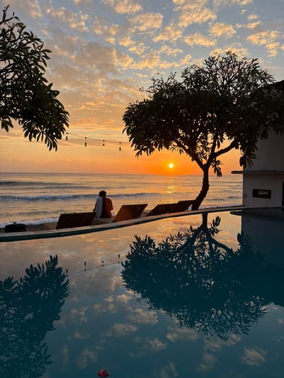 A tranquil sunset scene unfolds by the ocean in Abang, Indonesia. A lone figure, draped in a white and orange garment, sits facing the horizon, seated on a lounge chair near a swimming pool. The sun, a brilliant orb, dips towards the water, casting a warm, golden glow across the sky, which is adorned with scattered, wispy clouds. The gentle waves of the ocean create a soothing rhythm against the shore, which is lined with more lounge chairs. A tropical tree, silhouetted against the vibrant sky, stands to the right, its branches reaching outwards. The serene atmosphere is further enhanced by the still, reflective surface of the swimming pool, which mirrors the sunset, the sky, and the surrounding foliage, creating a breathtaking visual symmetry.