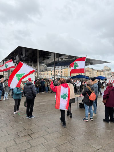 The image captures an outdoor gathering on a paved plaza under an overcast sky, identified as the Vieux-Port in Marseille, France, due to the prominent mirrored Ombrière structure visible in the background. Approximately 30-50 individuals are present, predominantly adults, dressed in various winter garments. Multiple participants are actively holding large Lebanese flags, distinguished by red horizontal stripes at the top and bottom, a white central band, and a green cedar tree in the middle. One woman in the immediate foreground, viewed from behind, wears a red jacket with a Lebanese flag draped over her back like a cape, holding a dark stick or pole in her right hand, gesturing towards the crowd. Another individual in the mid-ground wears a black puffer jacket and a red cap, holding a large Lebanese flag. A cluster of people congregates towards the mid-right background, near a small table displaying papers. A white sign with black text, partially obscured, shows phrases including "ARRETE Z..." and "UJE". The stone-block paved ground appears damp.
