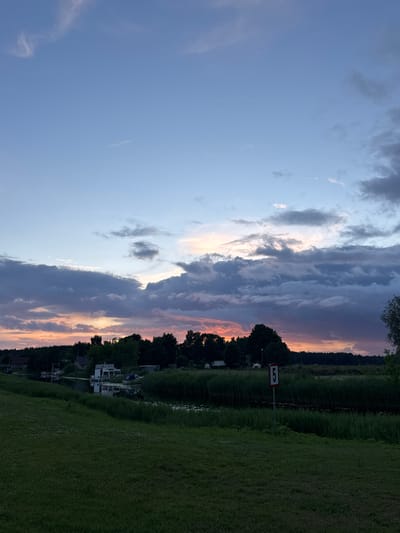 A wide-angle, low-light, exterior shot captures a sunset over the Gauja River in Carnikava, Latvia. The sky transitions from a pale blue at the top to hues of pink, orange, and deep purple near the horizon, with dramatic clouds creating a textured, layered effect. In the foreground, a dark green, manicured lawn slopes gently towards a dense embankment of reeds lining the river. The river itself is dark and reflective. Along the riverbank, a cluster of buildings and boats are visible. Notably, a white structure with an ornate lattice design, possibly a boathouse or small pavilion, stands out. Several boats are moored nearby. A red and white sign with the number "5" is planted in the grassy area, facing the river. The overall scene depicts a serene, twilight atmosphere in a waterside setting.